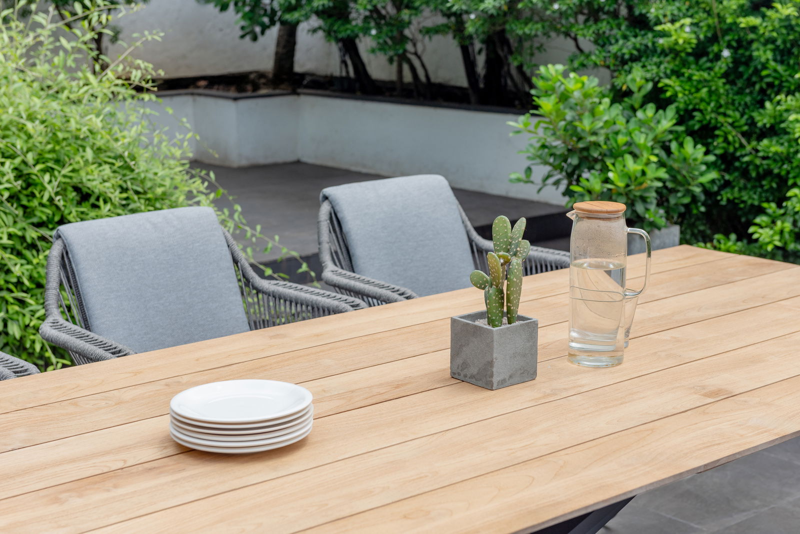 Gartentisch aus Holz mit grauen Stühlen, einem Kaktus in einem Beton-Topf und einer Glaskaraffe mit Wasser, aus der Perspektive von schräg oben fotografiert.