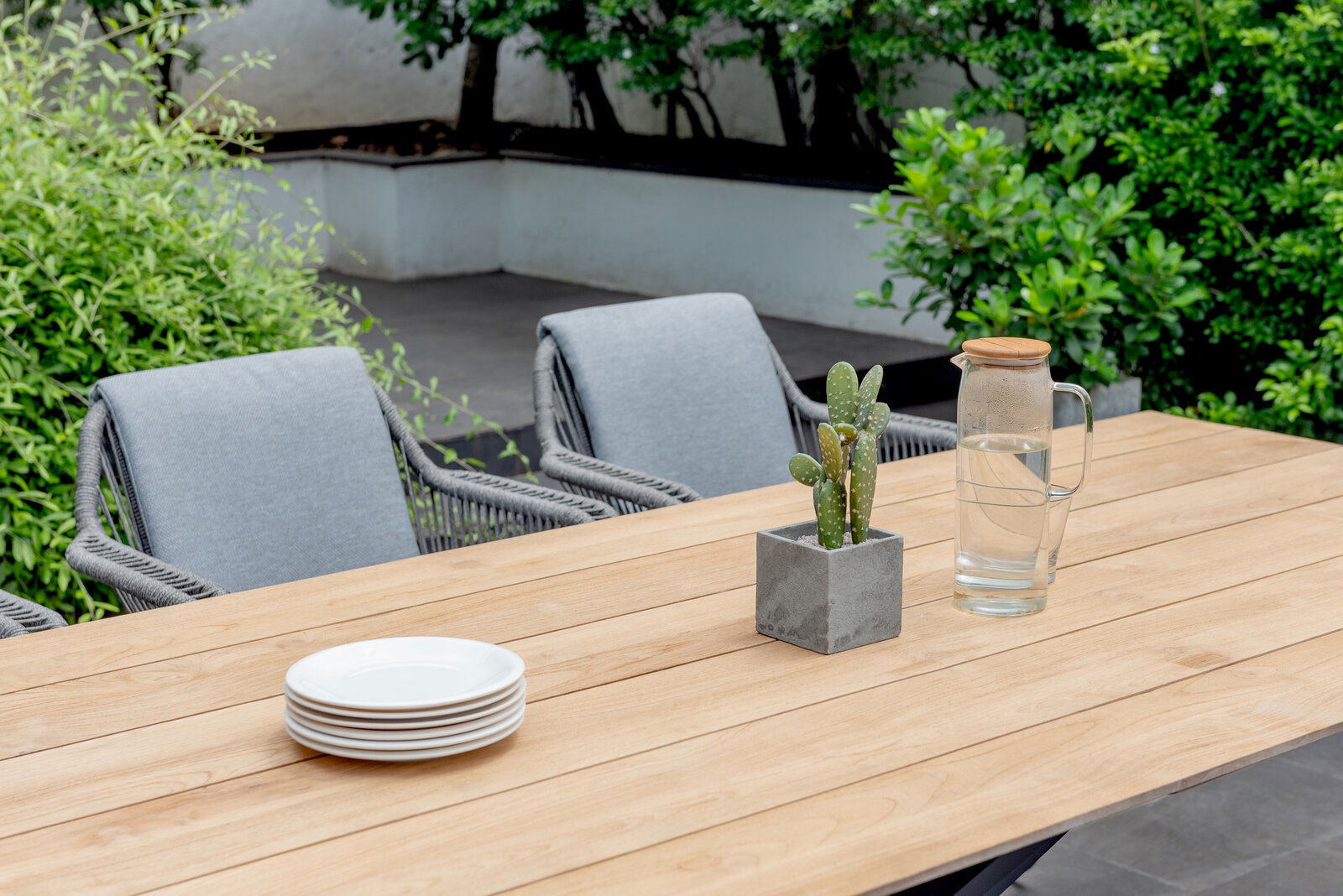 Gartentisch aus Holz mit grauen Stühlen, einem Kaktus in einem Beton-Topf und einer Glaskaraffe mit Wasser, aus der Perspektive von schräg oben fotografiert.