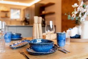 Modern gedeckter Esstisch mit blau glasiertem Keramikgeschirr, Gläsern und Holz-Salz-/Pfeffermühlen in einer warm beleuchteten Küche, aufgenommen aus leicht seitlicher Perspektive auf Tischhöhe.