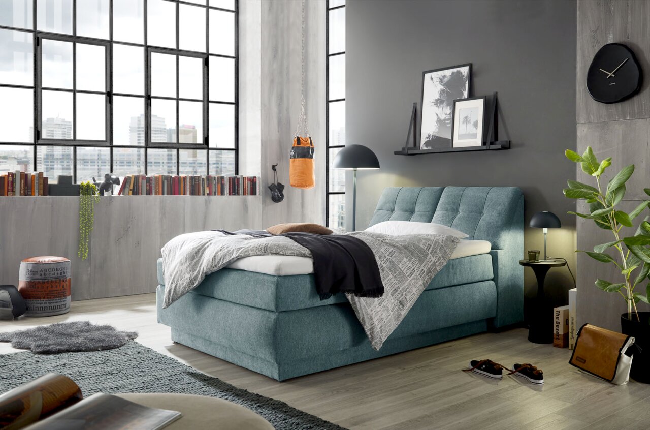 Boxspringbett in modernem Schlafzimmer mit großen Fenstern, grauer Wand und stilvoller Dekoration, seitliche Perspektive