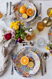 Draufsicht auf einen gedeckten Tisch mit buntem Patchwork-Porzellanservice (Teller und Schalen), Zitrusfrüchten, Gläsern, Besteck, Leinenservietten und Blumenstrauß auf hellem Untergrund