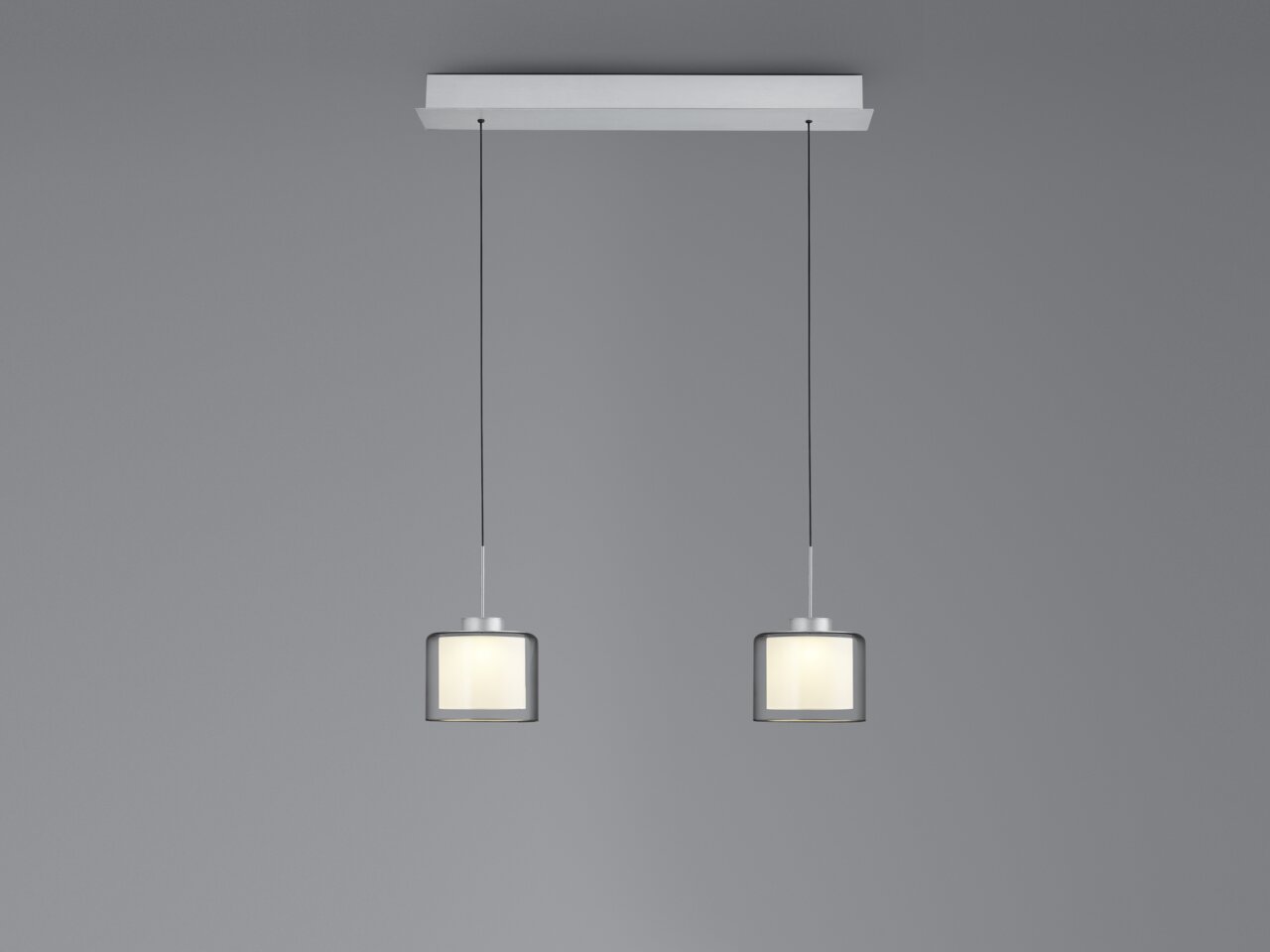 Moderne LED-hanglamp met twee cilindervormige lampenkappen, hangend aan dunne kabels aan een rechthoekige plafondbevestiging, zijaanzicht.