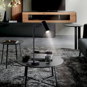 Moderne schwarze LED-Tischleuchte mit zylindrischem Leuchtenkopf auf rundem Sockel, auf einem runden Couchtisch im Wohnzimmer; Perspektive: frontale leicht erhöhte Ansicht