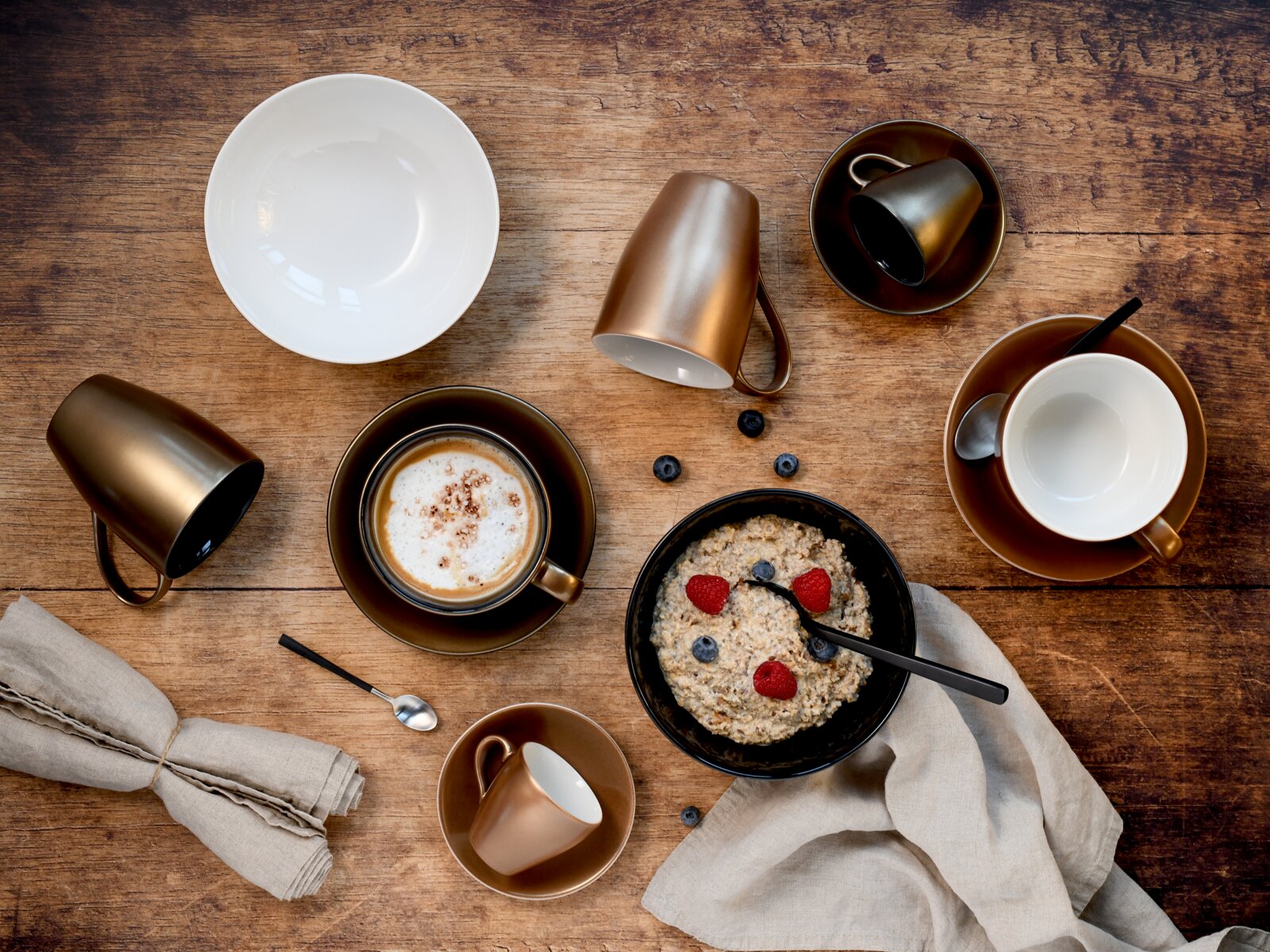 Draufsicht auf rustikalen Holztisch mit braunem Kaffeegeschirr (Tassen und Untertassen), einer Schale Porridge mit Himbeeren und Blaubeeren sowie Leinenservietten und Löffel.