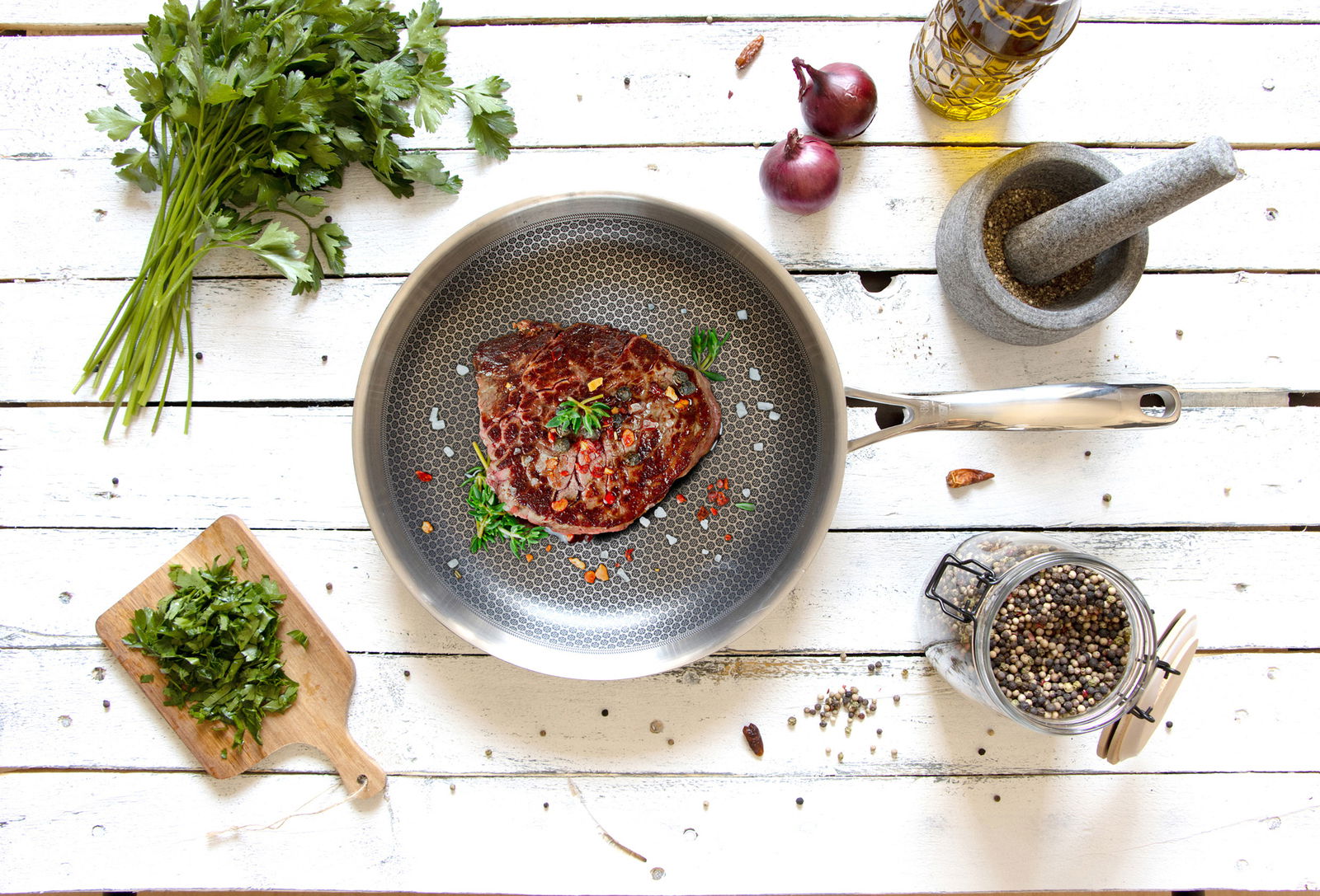 Bratpfanne RELIEF Evo 28x5,5cm mit gebratenem Steak, von oben fotografiert, umgeben von frischen Kräutern, Zwiebeln und Gewürzen auf einem weißen Holztisch.