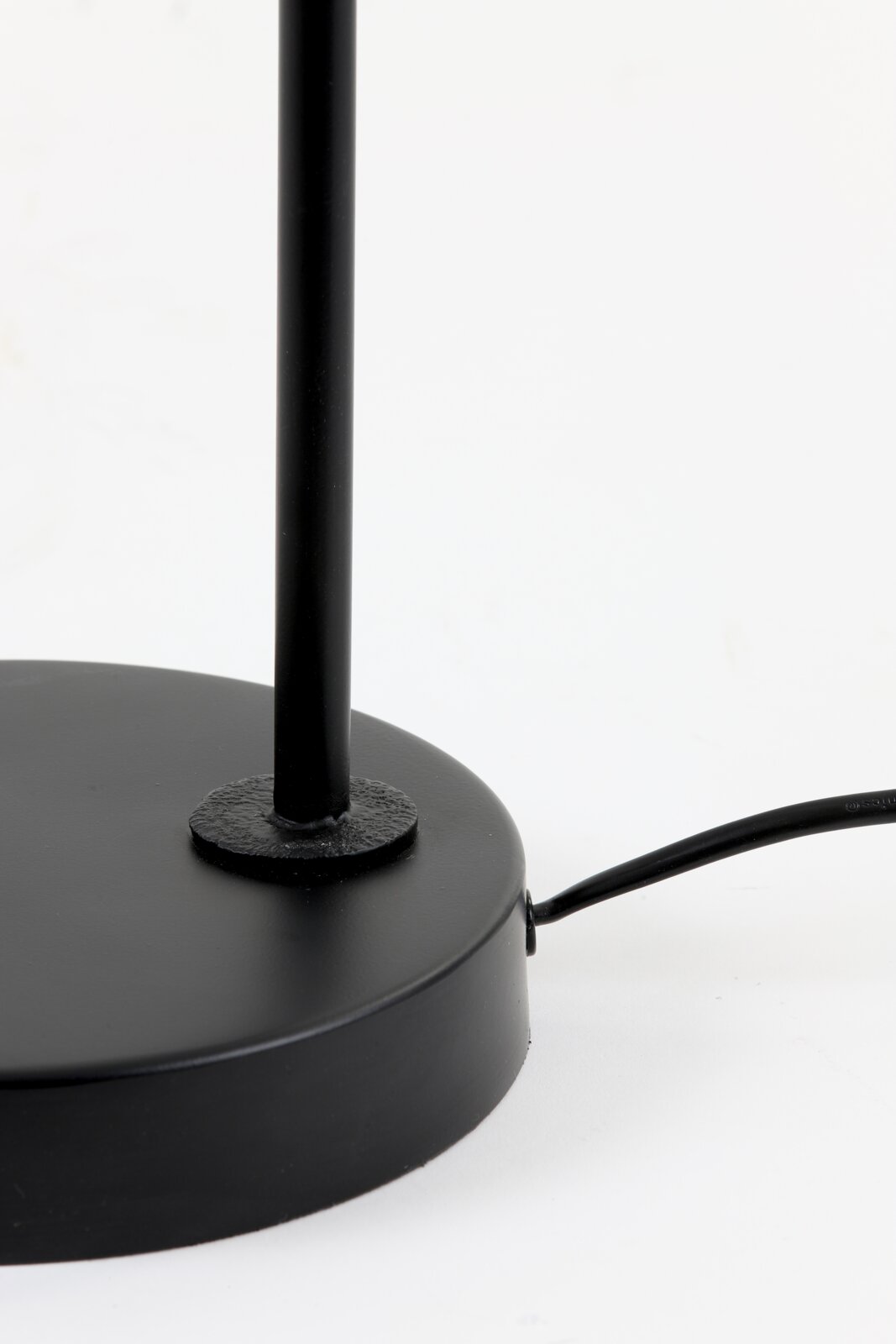 Close-up in vooraanzicht van de matzwarte ronde voet en slanke staander van een lamp, met zichtbaar zwart snoer op een witte achtergrond