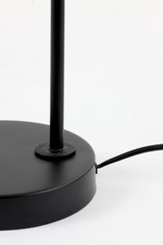 Close-up in vooraanzicht van de matzwarte ronde voet en slanke staander van een lamp, met zichtbaar zwart snoer op een witte achtergrond
