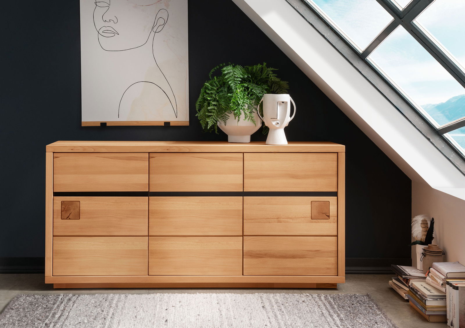Landscape Sideboard BOZEN Holz-Sideboard mit sechs Schubladen, dekoriert mit einer Pflanze und einer Vase, vor einer dunklen Wand und einem Dachfenster, aus frontaler Perspektive.
