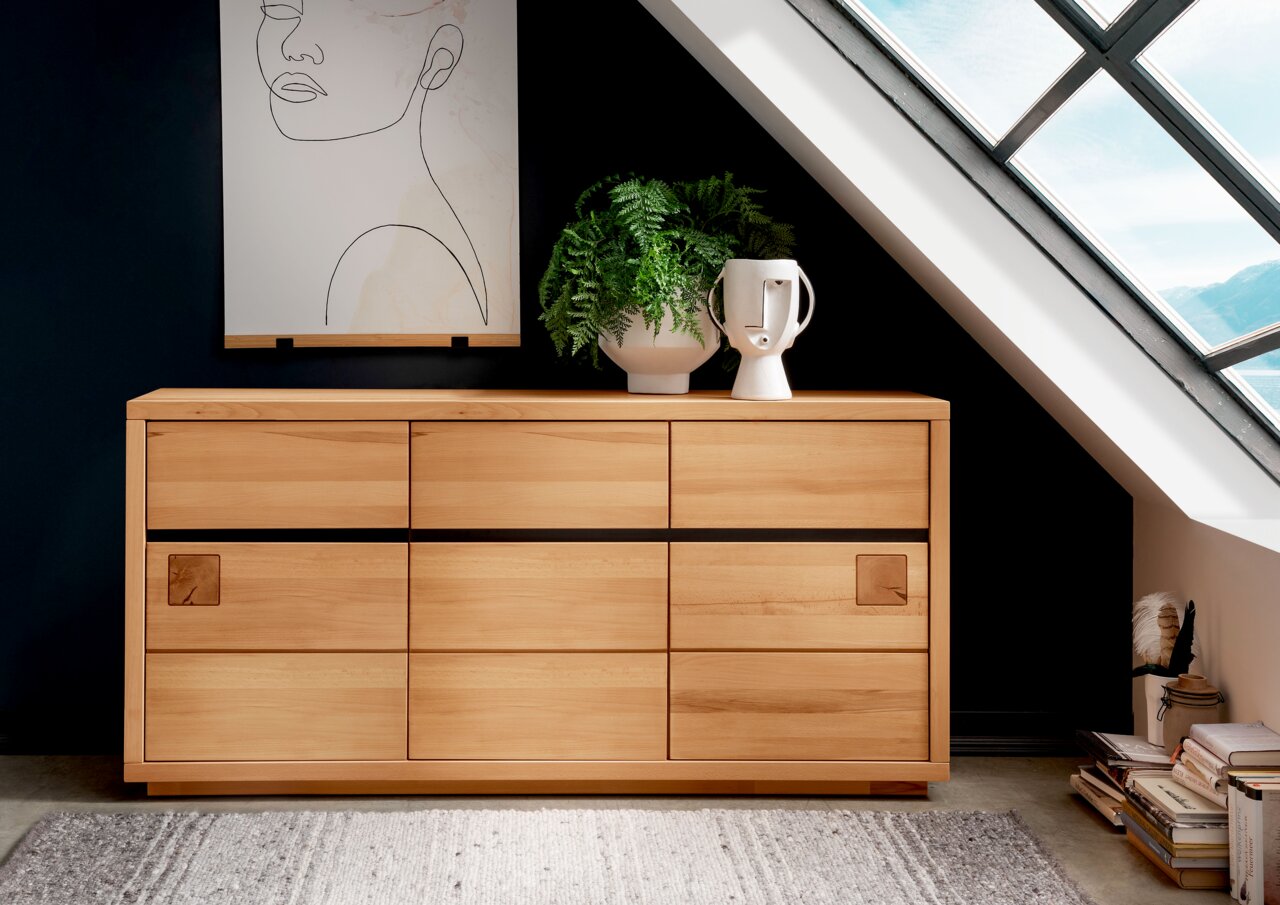 Landscape Dressoir BOZEN Houten dressoir met zes lades, versierd met een plant en een vaas, voor een donkere muur en een dakraam, gezien vanuit een vooraanzicht.