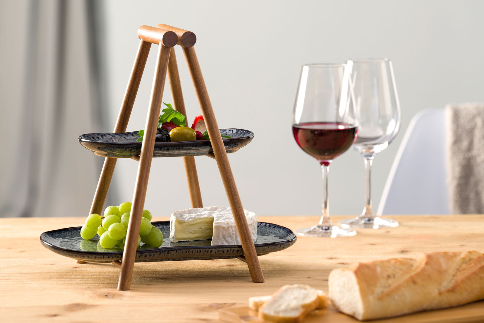 Leonardo Etagere 2 stöckig MATERA Zweistöckige Etagere aus Holz mit schwarzen Tellern, gefüllt mit Trauben, Käse und Oliven, auf einem Tisch mit Weingläsern und Baguette, seitliche Perspektive.
