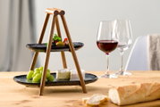 Zweistöckige Etagere aus Holz mit schwarzen Tellern, gefüllt mit Trauben, Käse und Oliven, auf einem Tisch mit Weingläsern und Baguette, seitliche Perspektive.