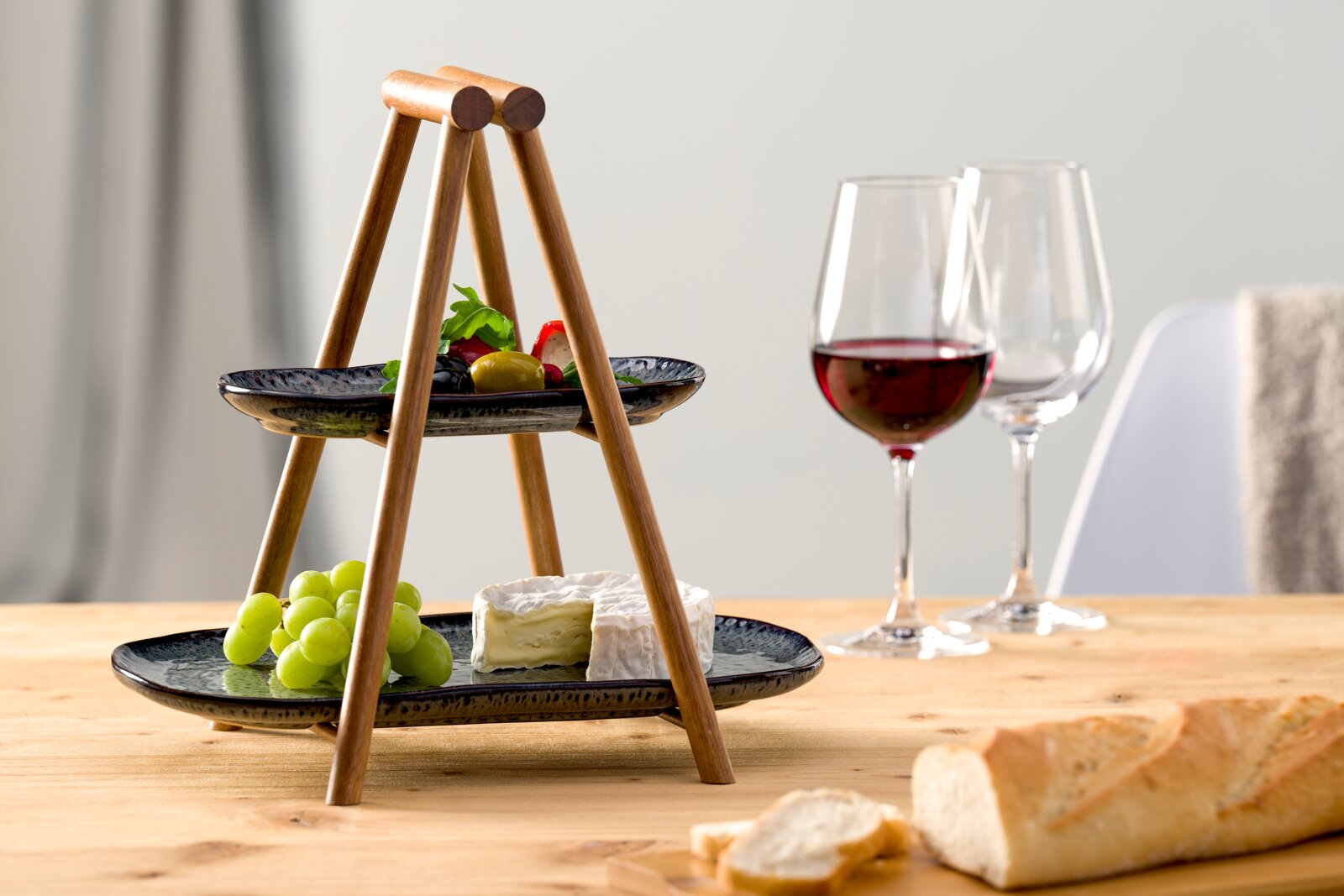 Houten etagère met twee verdiepingen en zwarte borden, gevuld met druiven, kaas en olijven, op een tafel met wijnglazen en stokbrood, zijaanzicht.