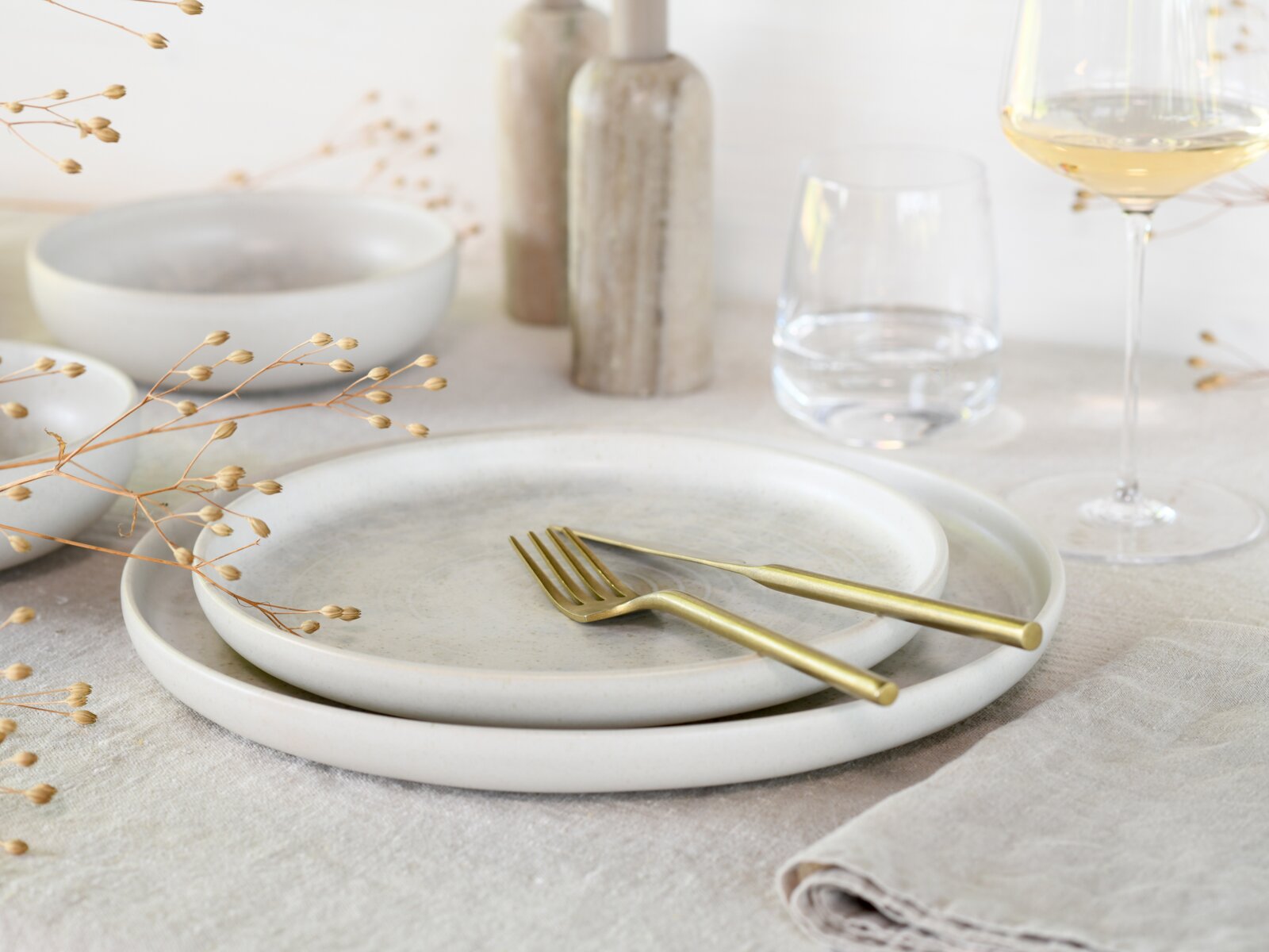 Minimalistisches Tisch-Setting mit zwei hellen Keramiktellern und goldfarbenem Besteck auf Leinentuch, seitliche Nahaufnahme in leichter Aufsicht, mit Weinglas, Wasserglas und Deko im Hintergrund
