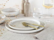 Minimalistisches Tisch-Setting mit zwei hellen Keramiktellern und goldfarbenem Besteck auf Leinentuch, seitliche Nahaufnahme in leichter Aufsicht, mit Weinglas, Wasserglas und Deko im Hintergrund