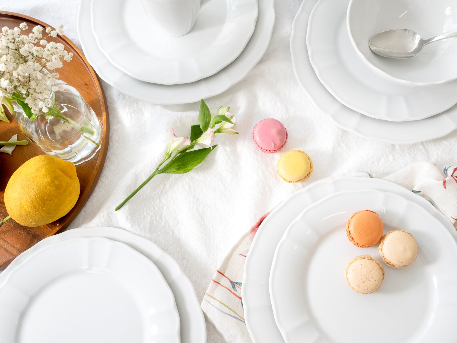 Draufsicht auf ein elegantes weißes Geschirr-Arrangement mit stapelbaren Tellern und Schalen auf weißer Tischdecke, dazu bunte Macarons, Zitrone, Glasvase mit kleinen weißen Blumen und ein Löffel.