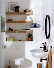 Frontale Raumansicht eines modernen, hellen Badezimmers mit weißem Wand-WC und Waschbecken, schwarzen Armaturen, ovalem Spiegel, Rauchglas-Pendelleuchte und dekorierten Holzregalen.