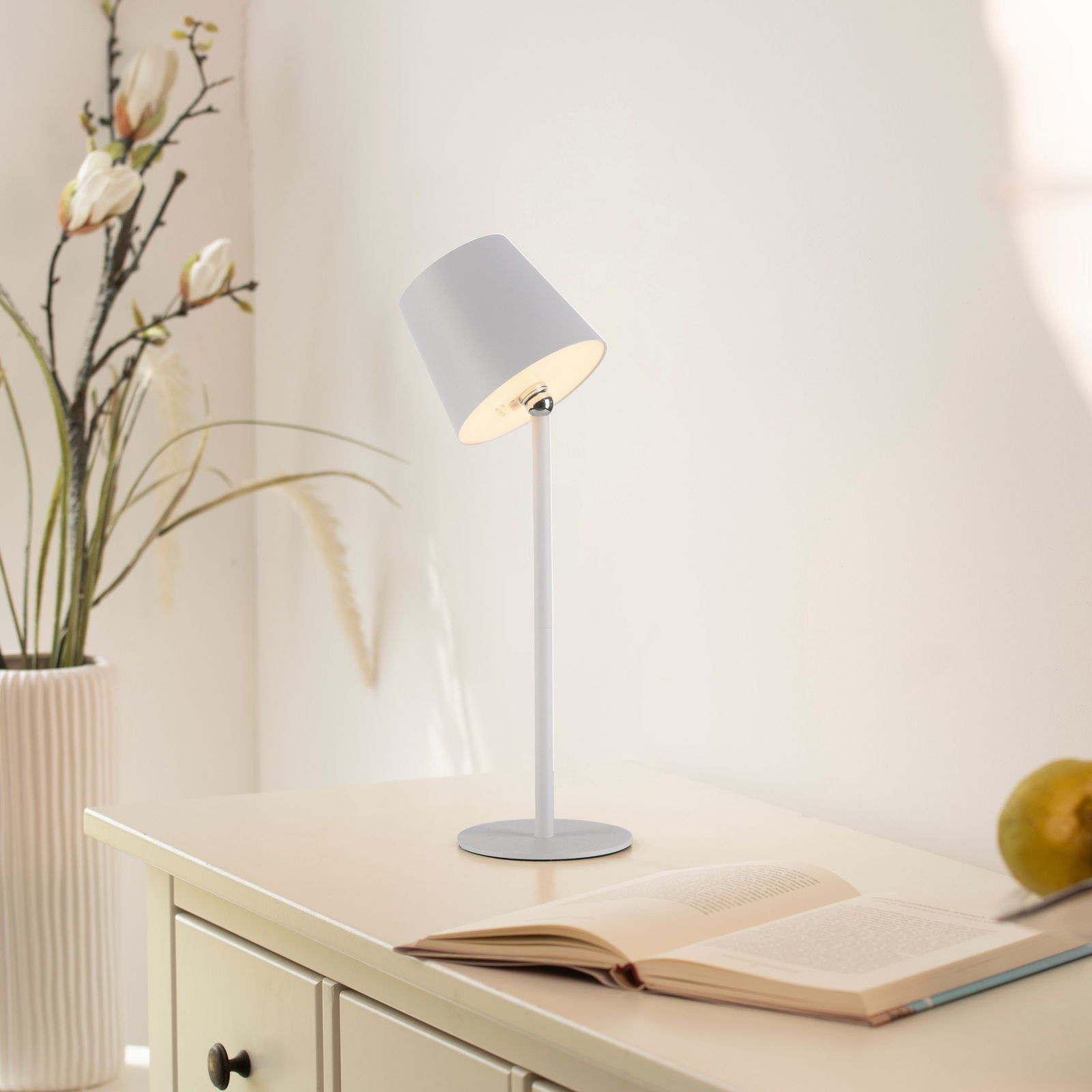 Moderne Akku-Tischleuchte BOTERIA in seitlicher Perspektive auf einem Tisch neben einem aufgeschlagenen Buch und einer Vase mit Blumen.