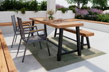 Moderner Gartentisch mit Holzplatte und schwarzen Metallbeinen, umgeben von zwei Stapelsesseln und einer Gartenbank, auf einer Terrasse mit Pflanzen im Hintergrund. Perspektive von der Seite.