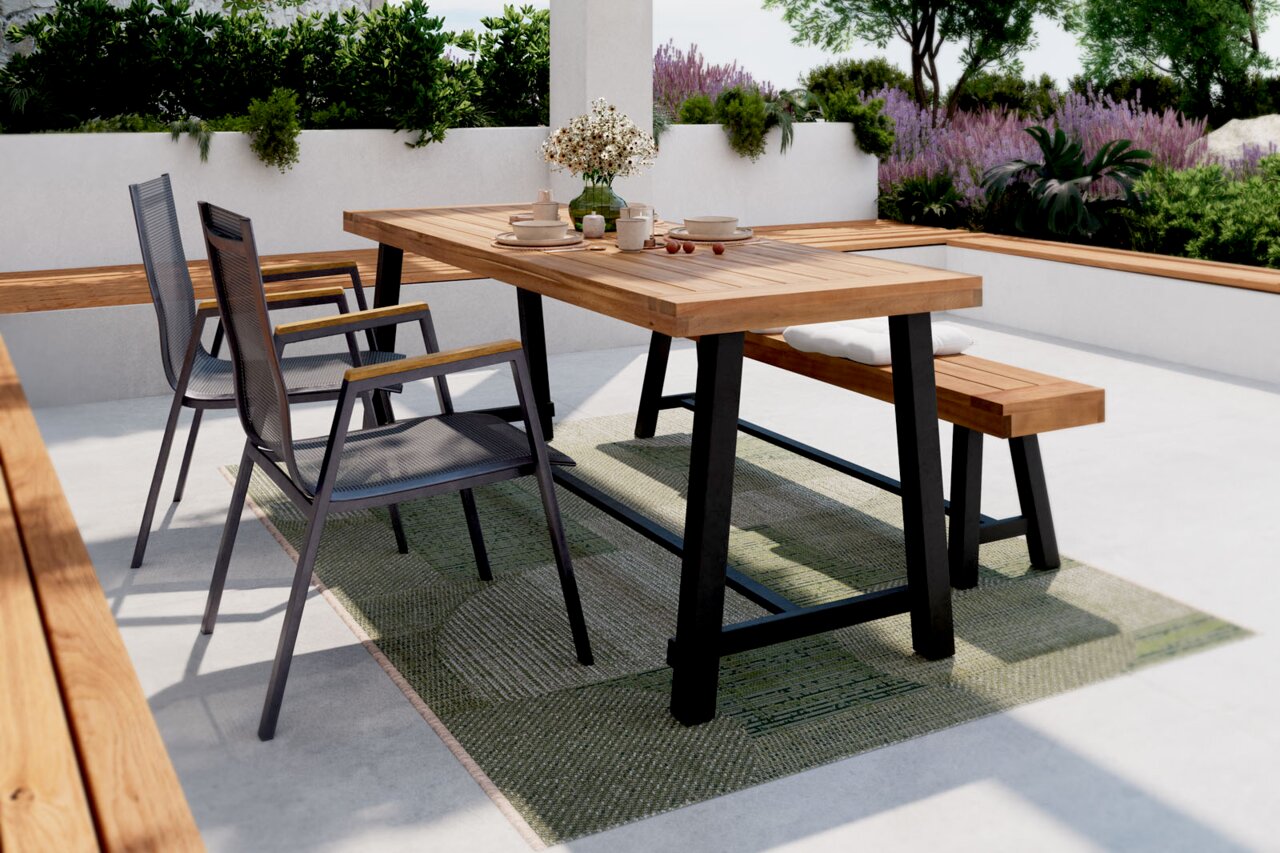 Moderner Gartentisch mit Holzplatte und schwarzen Metallbeinen, umgeben von zwei Stapelsesseln und einer Gartenbank, auf einer Terrasse mit Pflanzen im Hintergrund. Perspektive von der Seite.