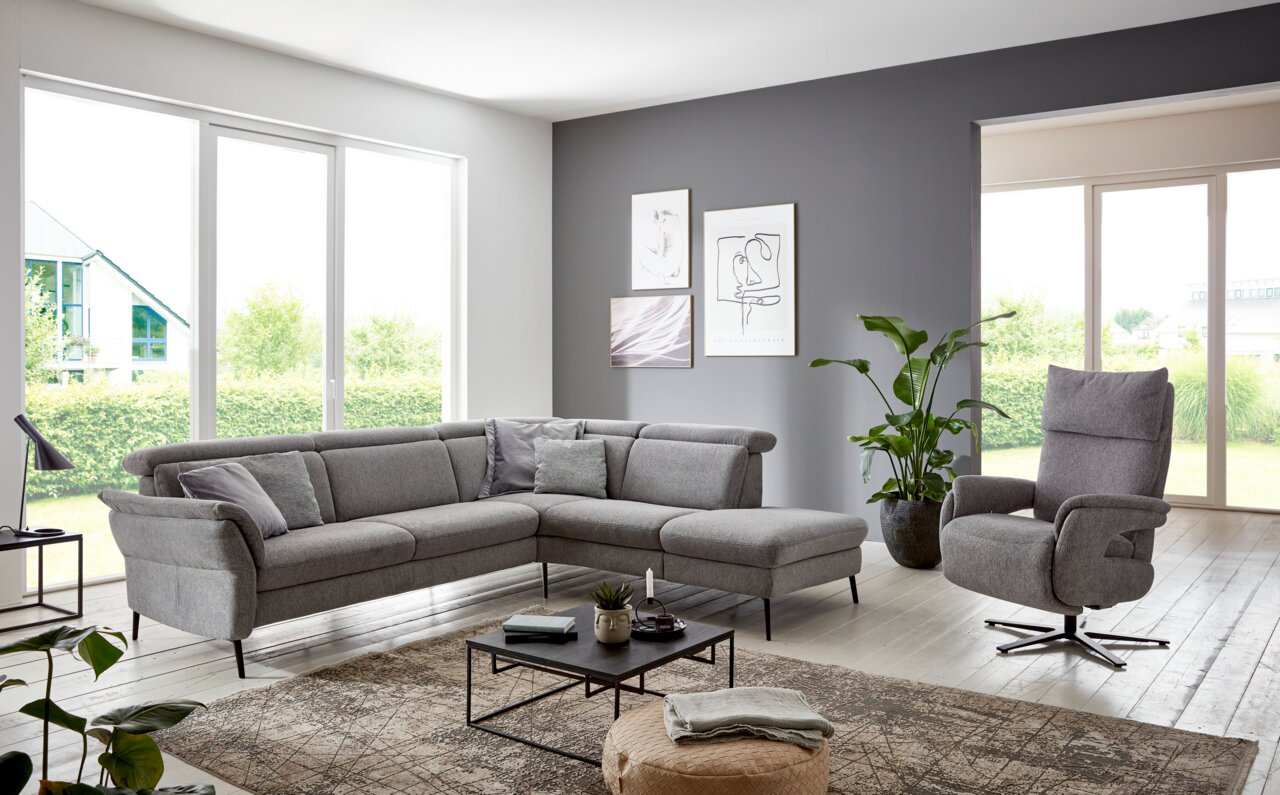 Graue Eckgarnitur in modernem Wohnzimmer mit großen Fenstern, Pflanzen und stilvollen Wandbildern, seitliche Perspektive.