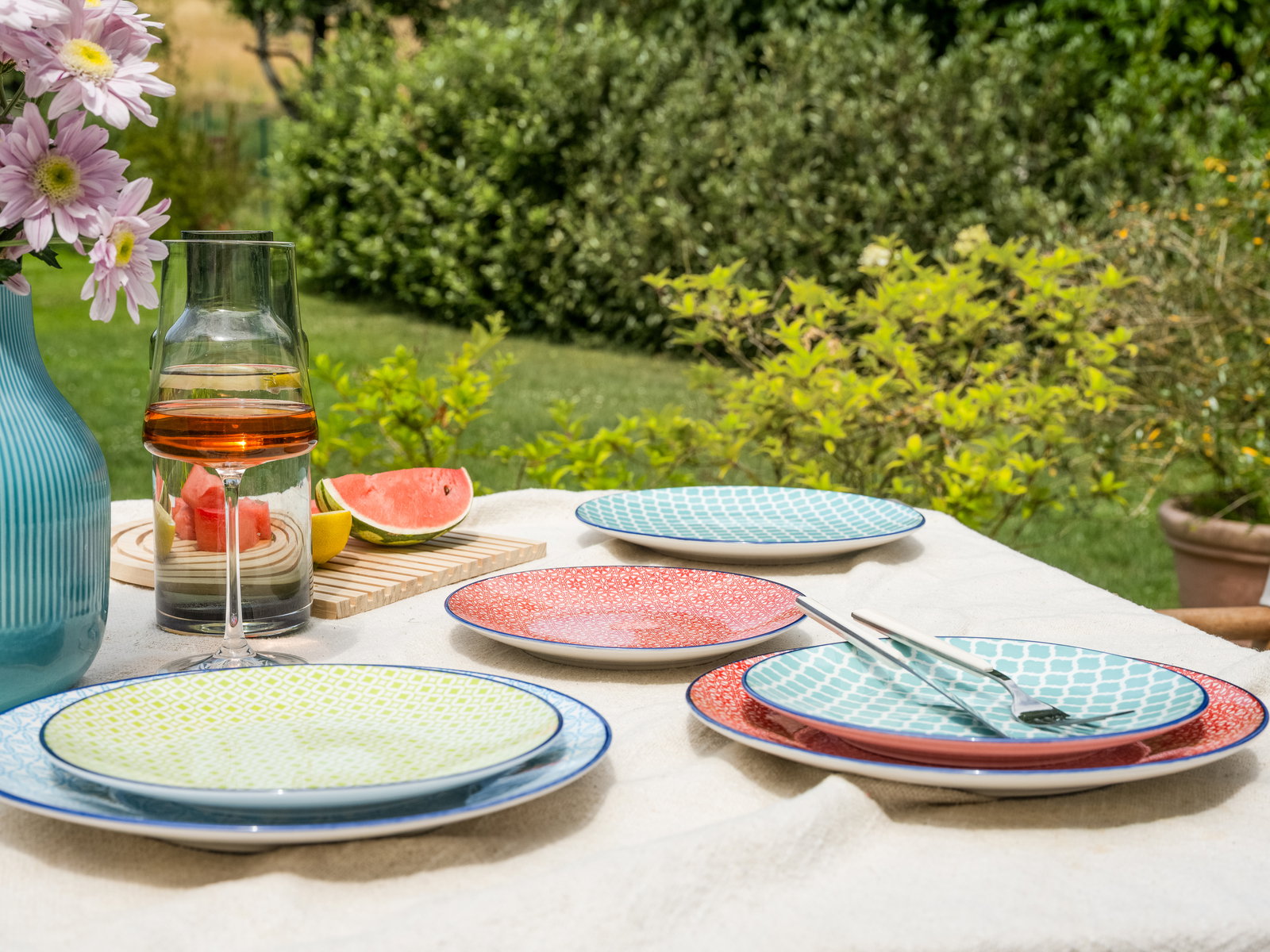 Ein Tellerset mit bunten Mustern auf einem Tisch im Garten, umgeben von Blumen und einem Getränkekrug, aus der Vogelperspektive fotografiert.