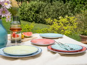 Ein Tellerset mit bunten Mustern auf einem Tisch im Garten, umgeben von Blumen und einem Getränkekrug, aus der Vogelperspektive fotografiert.