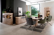 Modernes Esszimmer-Set mit großem Holzesstisch, gepolsterten Drehstühlen in Grau und Oliv sowie passenden Sideboards und Vitrine mit Beleuchtung, aufgenommen aus leicht seitlicher Perspektive auf Augenhöhe.