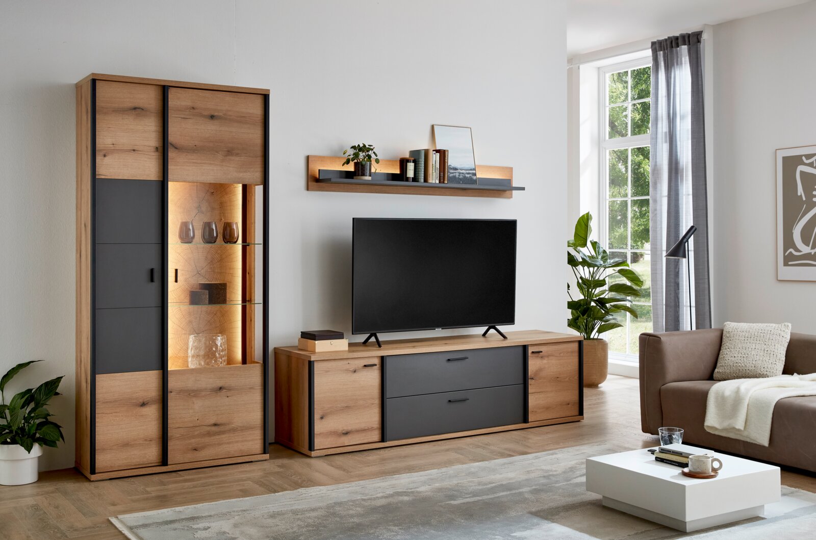 Wohnzimmer-Wohnwand im modernen Holz-und-Anthrazit-Design mit hoher Vitrine mit Beleuchtung, TV-Lowboard und Wandregal, frontal aus leicht seitlicher Perspektive fotografiert
