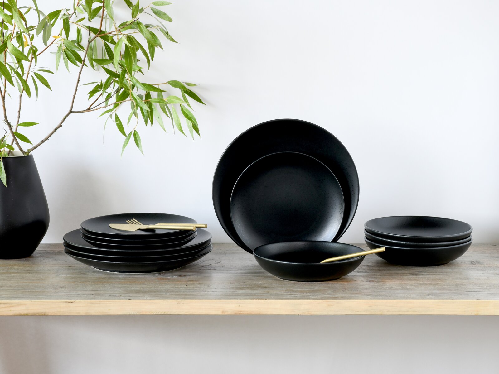 Creatable Tellerset 12-tlg MAGIC BLACK Matt schwarzes Geschirrset mit Tellern und Schalen auf einem Holztisch, dekoriert mit goldfarbenem Besteck und einer Vase mit grünen Zweigen; Ansicht leicht von vorne auf Augenhöhe