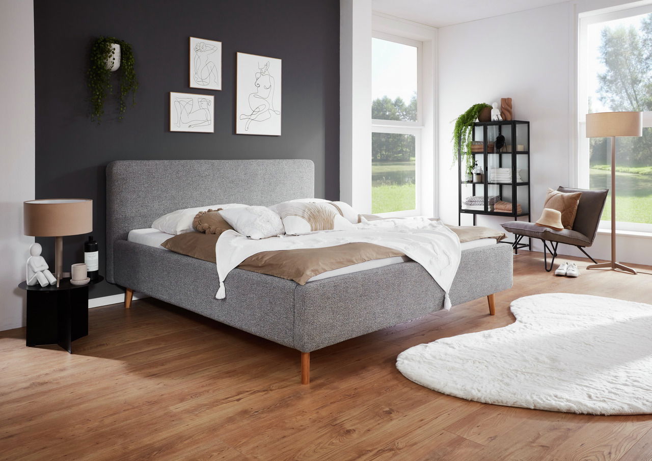 Modernes Polsterbettgestell in einem stilvoll eingerichteten Schlafzimmer mit grauem Kopfteil, Holzboden und großen Fenstern, seitliche Perspektive.