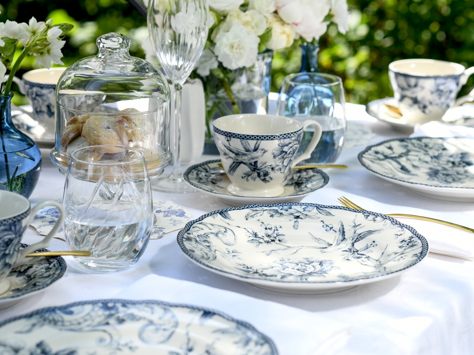 Schwarz-weißes Porzellan-Kaffeegeschirr mit floralen Vogelmotiven auf gedecktem Gartentisch, dazu Gläser, goldfarbenes Besteck und Kekse unter Glasglocke, aufgenommen aus leicht schräger Seitenperspektive auf Tischhöhe.