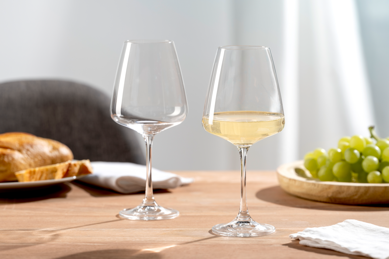 Leonardo Weißweinglas 6er-Set PALADINO Zwei elegante Weißweingläser auf einem Tisch, eines gefüllt mit Weißwein, im Hintergrund Trauben und Brot, seitliche Perspektive.