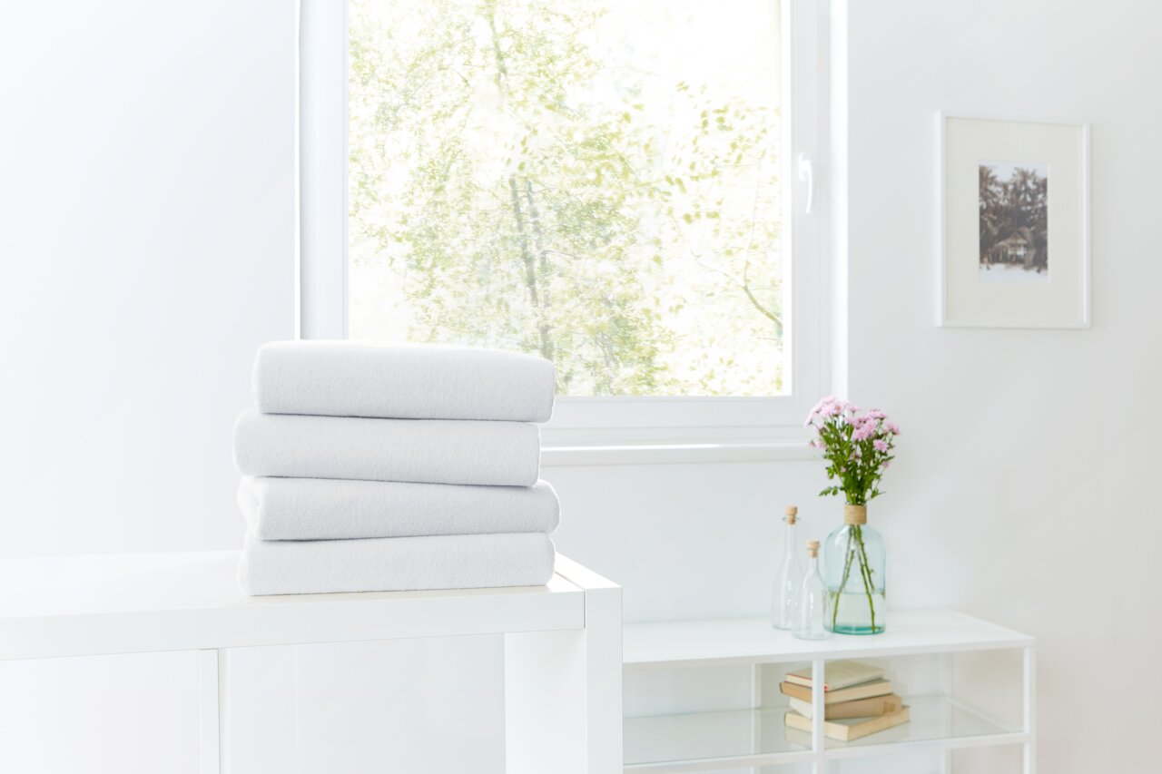 Novel Spannbetttuch TEDDY-FLAUSCH Ein Stapel weißer Spannbetttücher auf einem Tisch vor einem Fenster mit Blick auf einen hellen, grünen Garten.