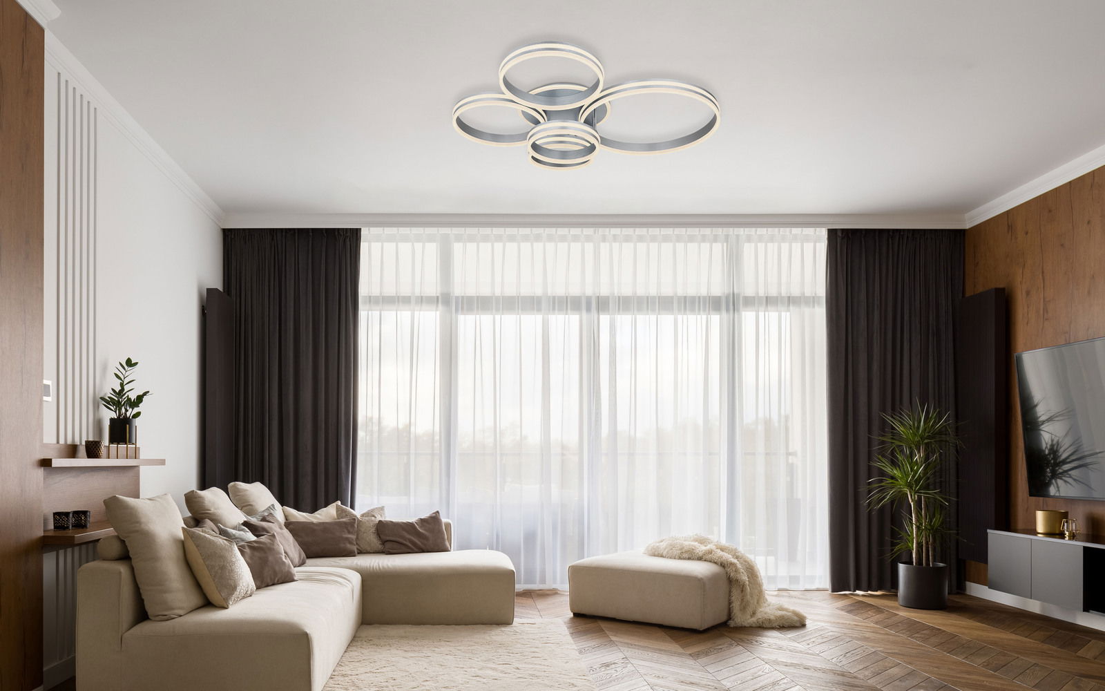 Moderne Deckenleuchte aus Metall in Grau, bestehend aus mehreren ineinander verschlungenen Ringen, von unten betrachtet in einem stilvollen Wohnzimmer mit großen Fenstern und eleganter Einrichtung.