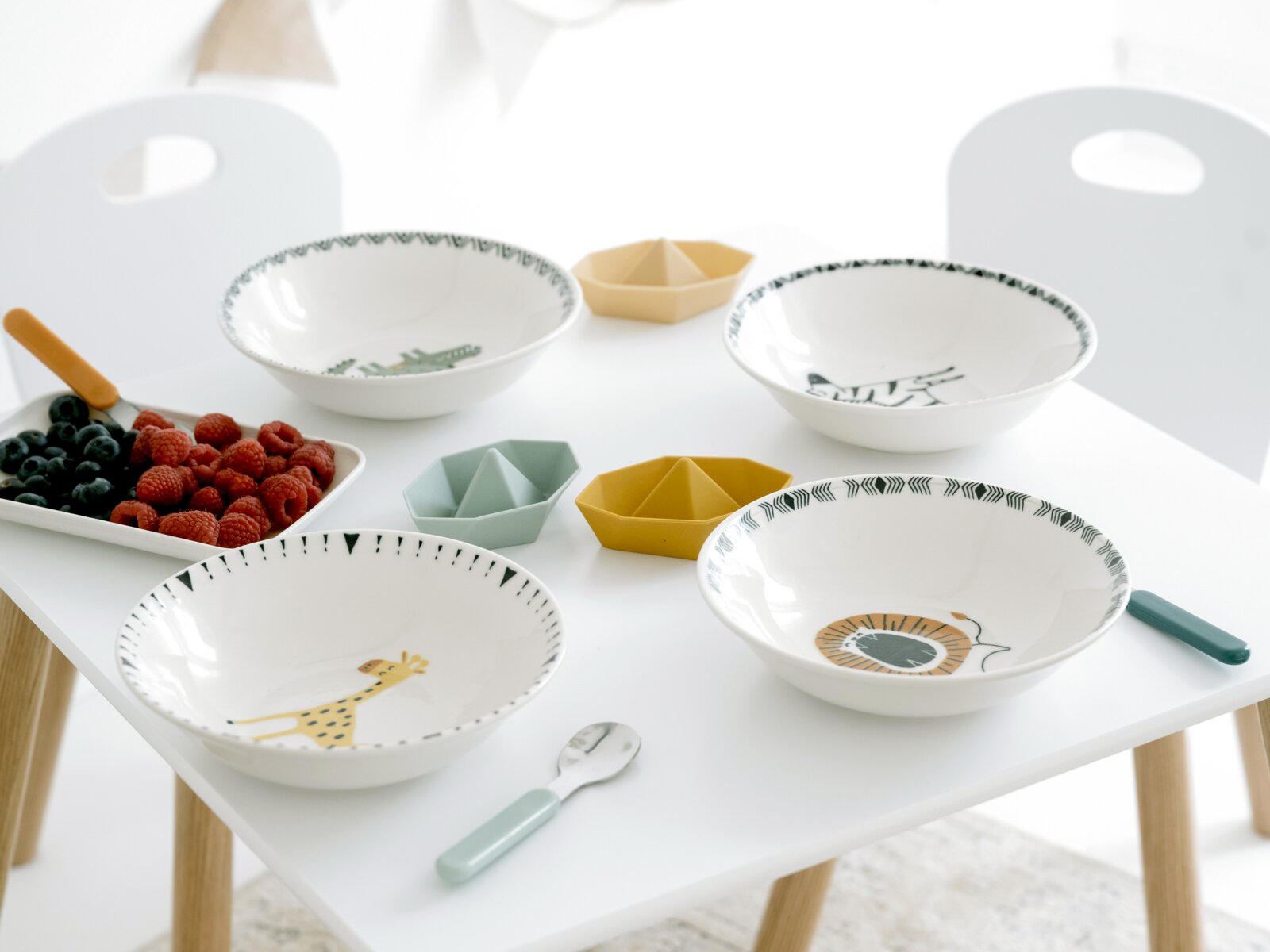 Schräg von oben fotografiertes Frühstücks-Set auf weißem Kindertisch: vier Keramikschalen mit Tiermotiven, bunte kleine Snackschalen, Löffel und Tablett mit Himbeeren und Blaubeeren.