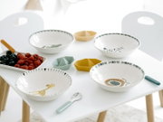 Schräg von oben fotografiertes Frühstücks-Set auf weißem Kindertisch: vier Keramikschalen mit Tiermotiven, bunte kleine Snackschalen, Löffel und Tablett mit Himbeeren und Blaubeeren.