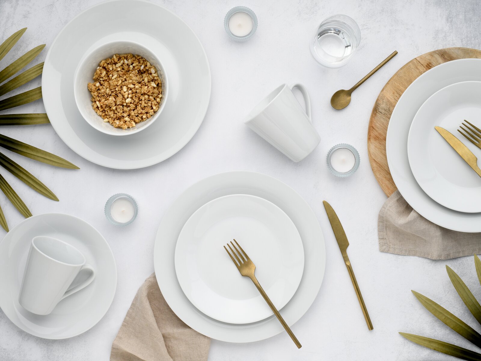 Draufsicht auf ein elegantes Tischgedeck mit weißem Porzellangeschirr (Teller, Schalen, Tassen), goldfarbenem Besteck und Schale mit Müsli auf hellem Hintergrund, ergänzt durch Kerzen, Wasserglas, Holzbrett und Leinentuch.