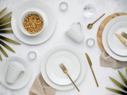 Draufsicht auf ein elegantes Tischgedeck mit weißem Porzellangeschirr (Teller, Schalen, Tassen), goldfarbenem Besteck und Schale mit Müsli auf hellem Hintergrund, ergänzt durch Kerzen, Wasserglas, Holzbrett und Leinentuch.