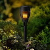 Solarbetriebene Gartenfackel aus schwarzem Plastik mit flackerndem Licht, in einem Garten aus der Seitenperspektive fotografiert.