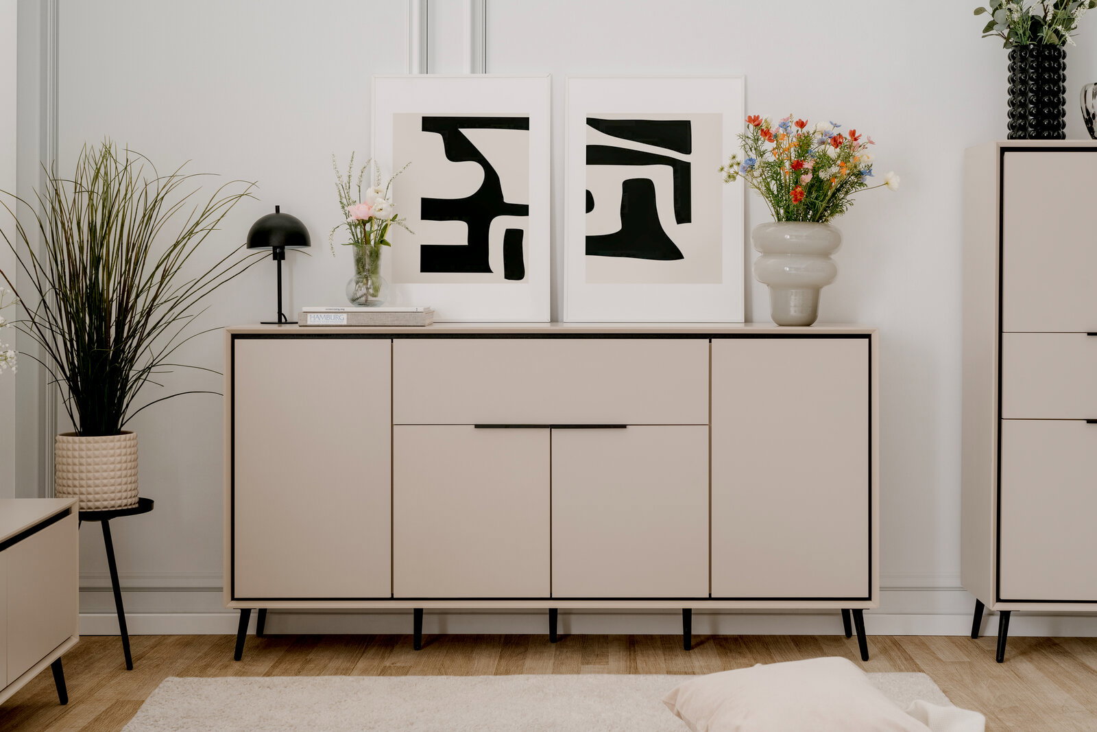 Modernes Sideboard in hellem Beige mit schwarzen Beinen, dekoriert mit Pflanzen und abstrakten Kunstwerken, Frontalansicht.