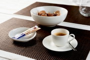 Weiße Espressotasse mit Untertasse und Löffel auf einem braunen Tischset, stilvoll arrangiert mit Zuckersticks und einer Schale Pralinen, aus leicht erhöhter schräger Perspektive fotografiert.