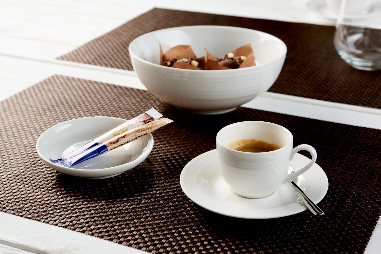 Weiße Espressotasse mit Untertasse und Löffel auf einem braunen Tischset, stilvoll arrangiert mit Zuckersticks und einer Schale Pralinen, aus leicht erhöhter schräger Perspektive fotografiert.