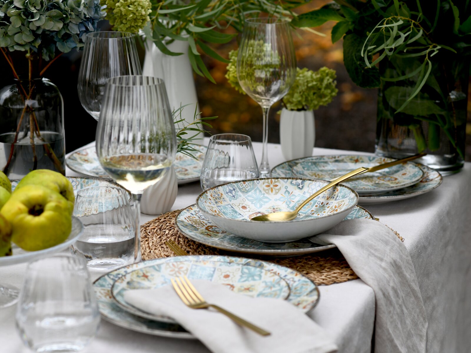 Schräg von oben fotografierte, stilvoll gedeckte Tafel mit gemustertem Keramikgeschirr in Blau- und Beigetönen, goldfarbenem Besteck, gerippten Weingläsern und Wassergläsern sowie grünen Blumenarrangements und Äpfeln.