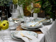 Schräg von oben fotografierte, stilvoll gedeckte Tafel mit gemustertem Keramikgeschirr in Blau- und Beigetönen, goldfarbenem Besteck, gerippten Weingläsern und Wassergläsern sowie grünen Blumenarrangements und Äpfeln.