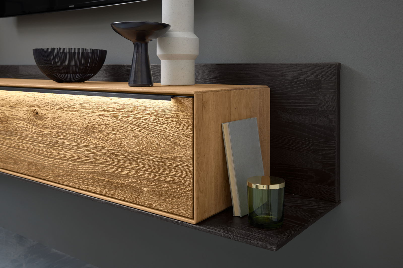 Nahaufnahme einer modernen Wandkombination mit Holzoptik und LED-Beleuchtung, dekoriert mit einer Vase, einer Schale und einem Buch, seitliche Perspektive.