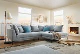 Moderne Eckgarnitur in einem hellen Wohnzimmer mit großen Fenstern, dekoriert mit blauen und grauen Kissen, aus der Perspektive von vorne links.