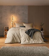 Gemütliches Schlafzimmer mit einem großen Bett in beiger Bettwäsche, dekoriert mit einem grauen Überwurf und Kissen, im Vordergrund ein flauschiger Teppich, seitliche Perspektive.