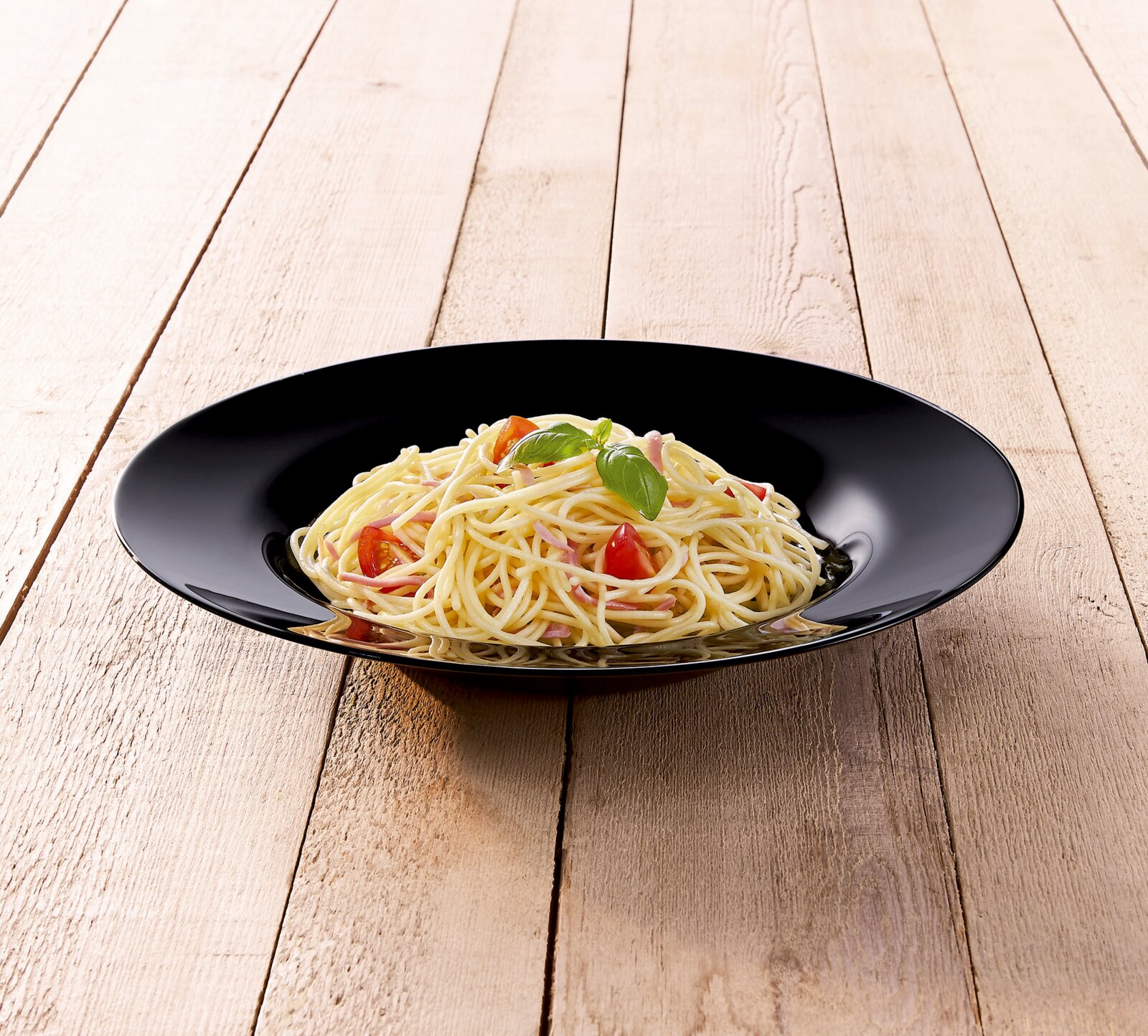 Schwarzer, tiefer Pastateller mit Spaghetti, Cherrytomaten, Schinkenstreifen und Basilikum auf hellem Holztisch, fotografiert aus leicht erhöhter Frontperspektive.