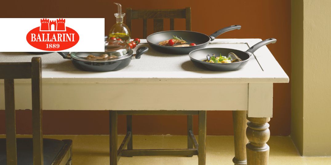 Op de foto is een rustieke keukentafel te zien met twee zwarte koekenpannen en het Ballarini-logo.