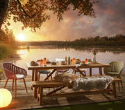 Gartenbank aus Holz auf einer Terrasse mit Blick auf einen See bei Sonnenuntergang, umgeben von Stühlen und einem gedeckten Tisch.